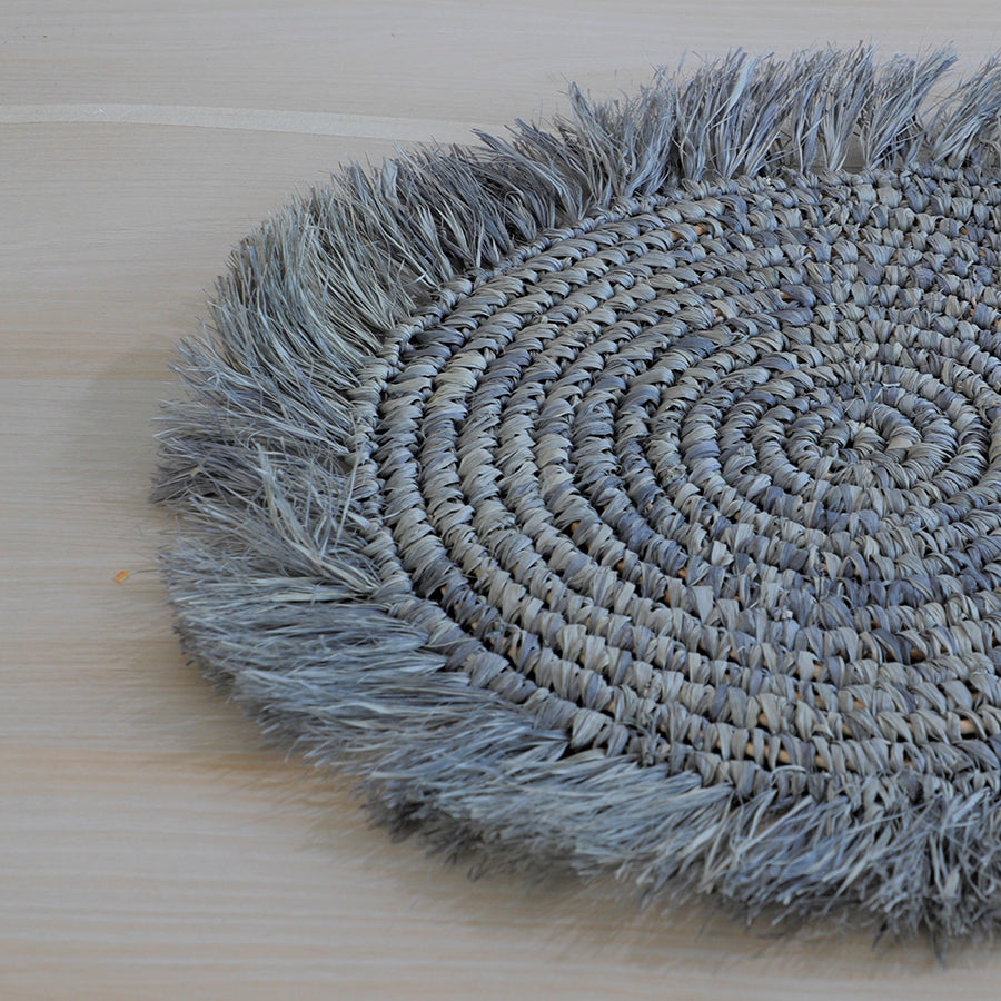 Grey woven fringe placemat, Boho round table mat, Handcrafted braided placemat, Natural straw dining mat, 
Jute-look table décor, Rustic table setting accessory, Textured bohemian placemat, Eco-friendly woven charger, Handmade round placemat with fringe, Spiral woven placemat grey. 