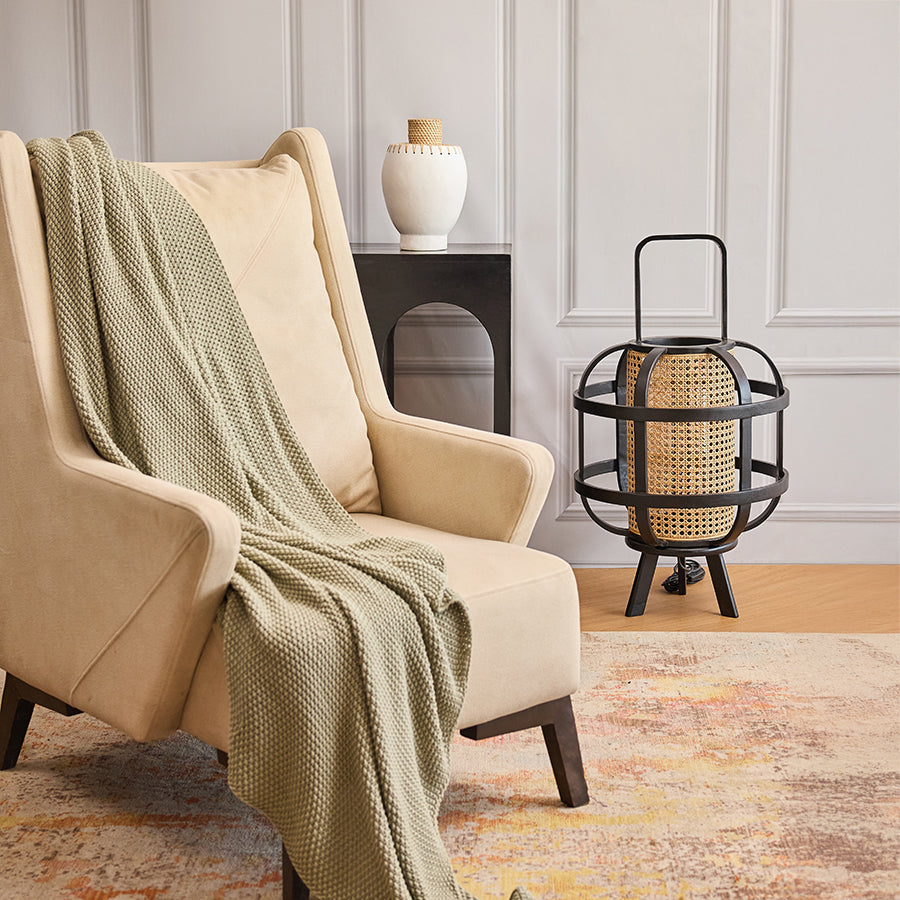 Cozy modern living room décor, Neutral tone interior styling, Mid-century modern beige accent chair,
Wingback armchair with wooden legs, Soft knitted sage green throw,
Cozy textured couch blanket, Handmade white ceramic vase,
Boho rattan neck vase, Black modern minimalist side table, Arched cutout accent table, Industrial style rattan lantern, Black cage lantern with woven detail, Abstract neutral color area rug, Vintage style living room rug.

